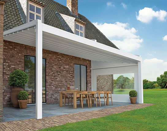 Modern white lean-to pergola attached to a brick house, covering an outdoor dining area with table and chairs in a garden setting