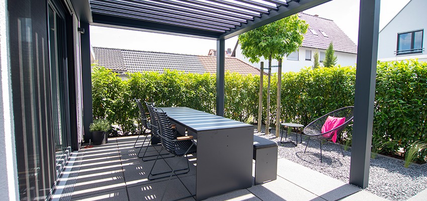 Modern aluminium pergola providing shaded outdoor dining area with sleek grey furniture and privacy hedging in a contemporary garden.